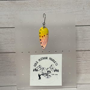 Puede incluir: Un señuelo de pesca con la parte superior amarilla, que pasa a naranja y rosa, y puntos negros. Tiene un anzuelo plateado y se muestra en una tarjeta de plástico transparente. La tarjeta tiene una pegatina con el texto "BDLR OUTDOOR PRODUCTS ESTD 2024".