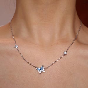 May include: A silver-toned necklace featuring a central butterfly pendant adorned with small blue and clear stones. The delicate chain includes two additional small accents, one star-shaped and one circular, both set with clear stones along the chain.