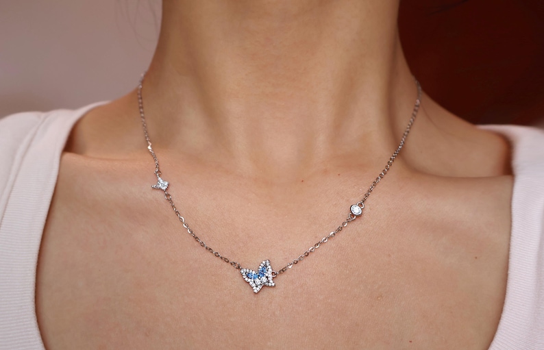 May include: A silver-toned necklace featuring a central butterfly pendant adorned with small blue and clear stones. The delicate chain includes two additional small accents, one star-shaped and one circular, both set with clear stones along the chain.