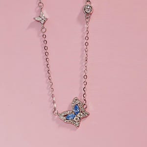 May include: A silver-toned chain necklace featuring a central butterfly pendant adorned with blue marquise-cut stones and clear pav&eacute; crystals. The chain includes a small star-shaped charm with clear crystals and a round bezel-set crystal accent.