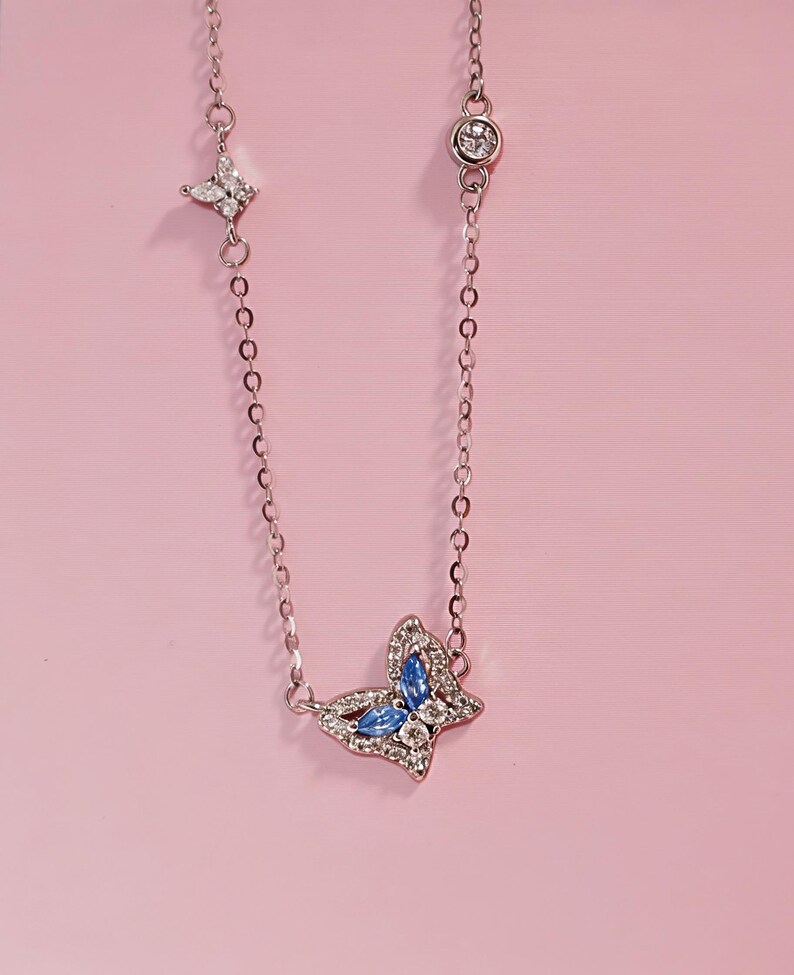May include: A silver-toned chain necklace featuring a central butterfly pendant adorned with blue marquise-cut stones and clear pav&eacute; crystals. The chain includes a small star-shaped charm with clear crystals and a round bezel-set crystal accent.