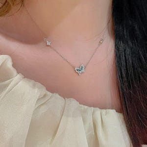 May include: A delicate silver-toned necklace featuring a central butterfly pendant adorned with small clear and blue stones. The chain includes a smaller butterfly accent on the left side, with a thin, polished metal link design throughout the piece.
