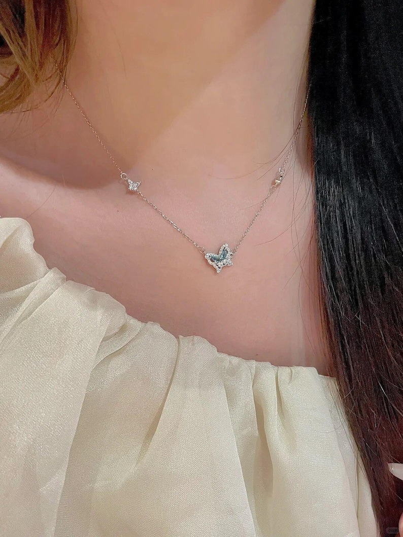 May include: A delicate silver-toned necklace featuring a central butterfly pendant adorned with small clear and blue stones. The chain includes a smaller butterfly accent on the left side, with a thin, polished metal link design throughout the piece.