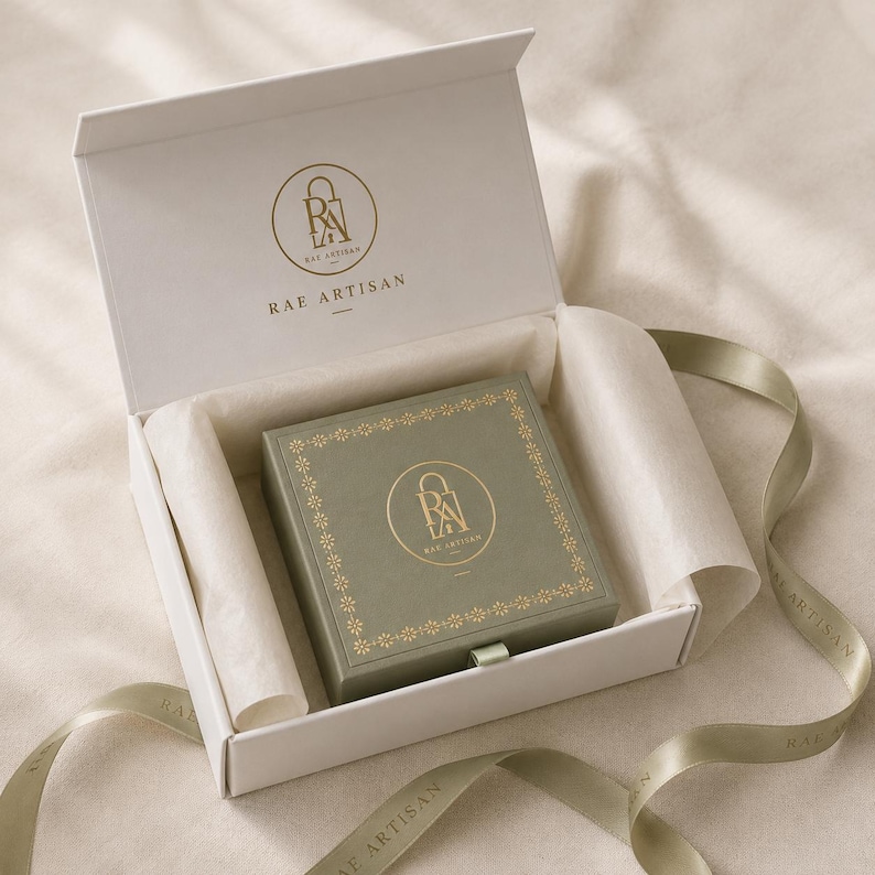 May include: A white hinged gift box containing a sage green square box with a gold floral border and the Rae Artisan logo. The interior lid features the same gold logo. A sage green ribbon with the Rae Artisan text is wrapped around the box.