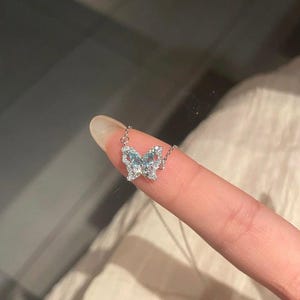 May include: A silver-toned necklace featuring a butterfly-shaped pendant. The butterfly wings are encrusted with small, clear, sparkling stones and accented with light blue stones in the center, attached to a delicate silver-toned chain.