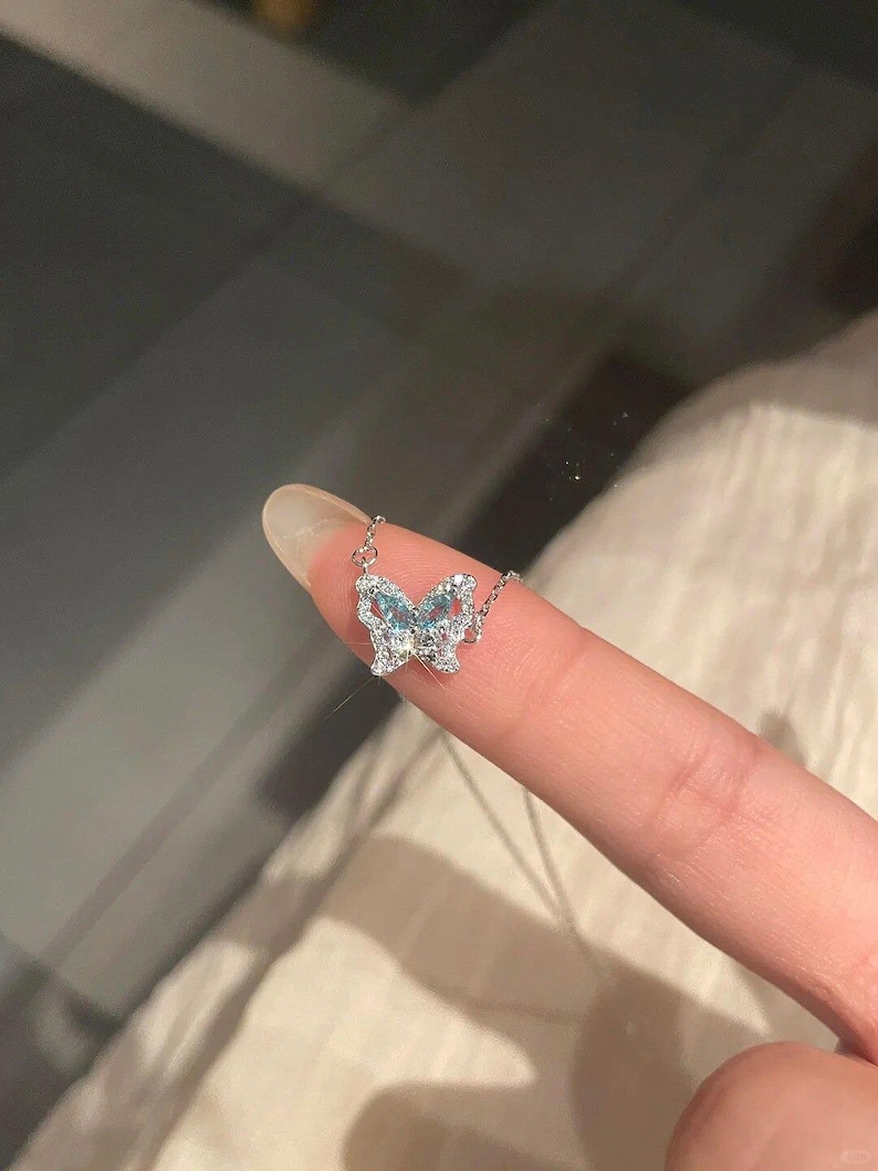 May include: A silver-toned necklace featuring a butterfly-shaped pendant. The butterfly wings are encrusted with small, clear, sparkling stones and accented with light blue stones in the center, attached to a delicate silver-toned chain.
