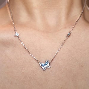 May include: A delicate silver-toned chain necklace featuring a central butterfly pendant adorned with small clear crystals and blue marquise-cut stones. The chain includes additional small decorative accents, including a tiny star-shaped charm.