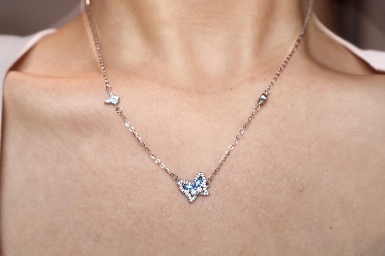 May include: A delicate silver-toned chain necklace featuring a central butterfly pendant adorned with small clear crystals and blue marquise-cut stones. The chain includes additional small decorative accents, including a tiny star-shaped charm.
