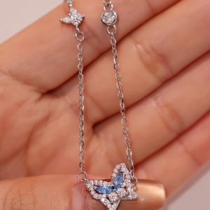 May include: A silver-toned chain bracelet featuring a butterfly-shaped charm adorned with small clear crystals and two marquise-cut light blue stones. The chain includes a small butterfly accent and a single round bezel-set clear crystal detail.