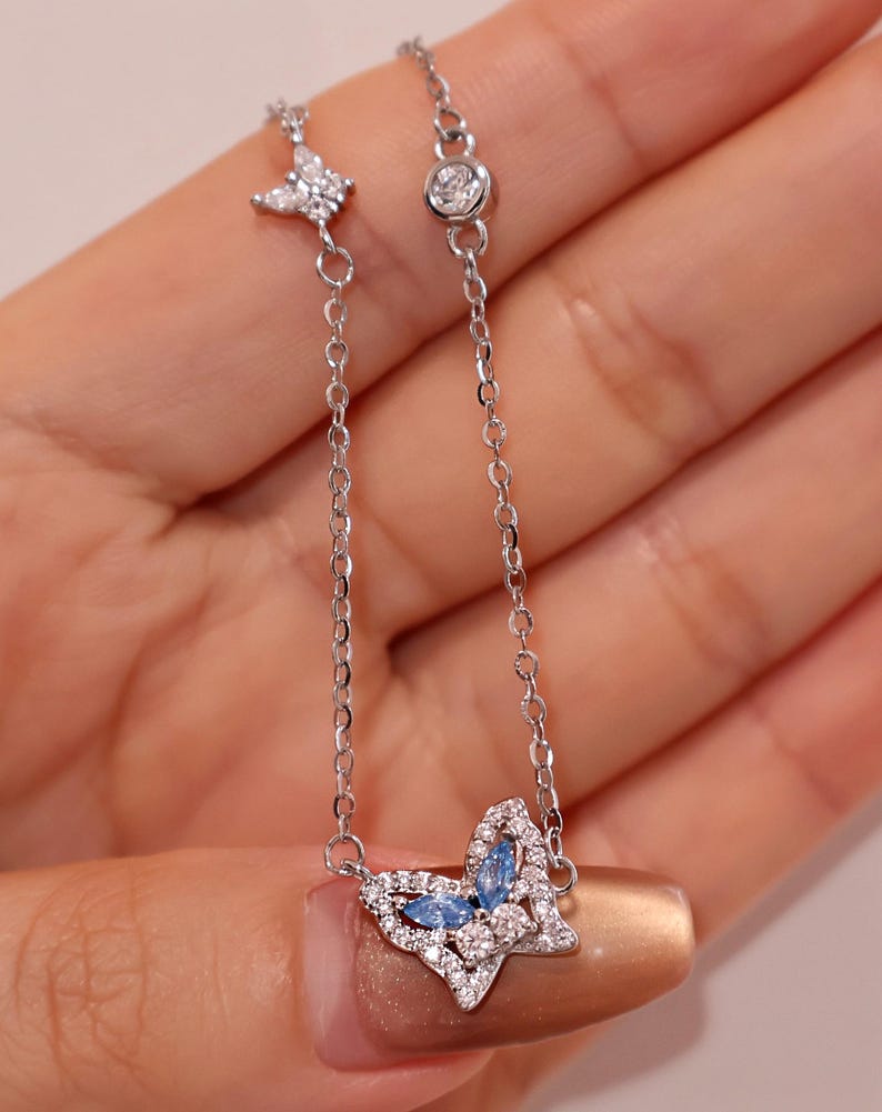 May include: A silver-toned chain bracelet featuring a butterfly-shaped charm adorned with small clear crystals and two marquise-cut light blue stones. The chain includes a small butterfly accent and a single round bezel-set clear crystal detail.