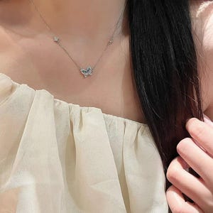 May include: A silver-toned necklace featuring a central butterfly pendant adorned with small, clear, sparkling stones. The delicate chain includes two additional small, stone-encrusted accents positioned symmetrically on either side of the butterfly.