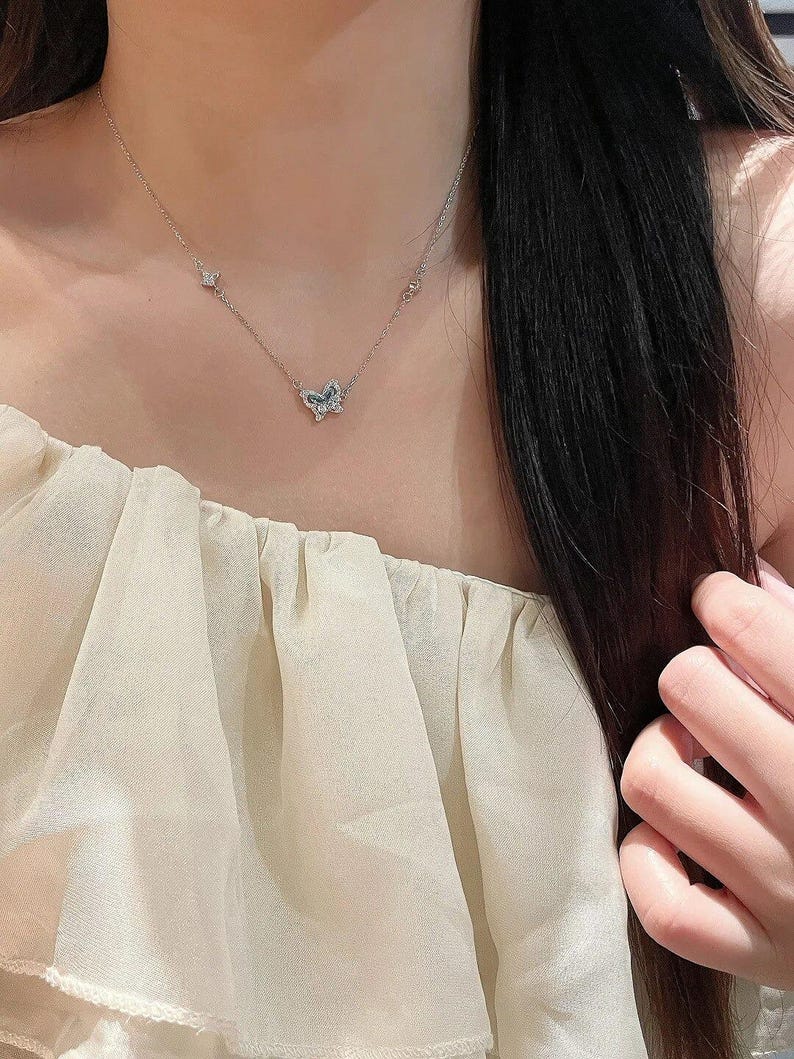 May include: A silver-toned necklace featuring a central butterfly pendant adorned with small, clear, sparkling stones. The delicate chain includes two additional small, stone-encrusted accents positioned symmetrically on either side of the butterfly.