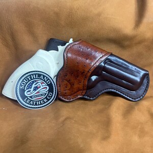 May include: A brown leather holster with a textured ostrich-style pattern on the outer panel and a smooth dark brown finish on the main body. A circular sticker with the text SOUTHLAND LEATHER CO BENTON AR and a cowboy hat graphic is included.