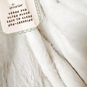May include: Close-up of a white, plush vegan fur blanket. The blanket has a soft, textured appearance. A tag in the upper left corner reads "interior" and lists features: "VEGAN FUR, ULTRA PLUSH, EASY TO CLEAN, NON-SHEDDING."