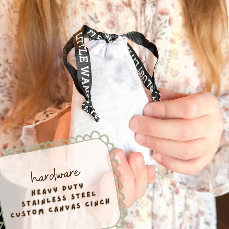 May include: A white canvas cinch bag with a black drawstring, held by a person. The drawstring has the text "LITTLE WANDERLUST" printed on it. The bag is labeled "hardware", "HEAVY DUTY", "STAINLESS STEEL", and "CUSTOM CANVAS CINCH".