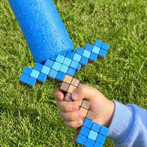May include: A blue foam sword with a pixelated design. The handle and crossguard feature a mosaic of blue and brown squares, while the blade is a solid blue cylinder. The toy is held against a backdrop of green grass.