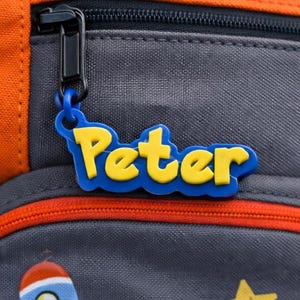 May include: Orange and gray backpack with space-themed patches, including a rocket, planet, and stars. A blue and yellow name tag that says "Peter" is attached to the zipper. The backpack has a mesh side pocket.