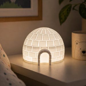 Puede incluir: Una luz de noche blanca en forma de iglú con un patrón de ladrillos rectangulares y una puerta curva. La lámpara está iluminada, emitiendo un brillo cálido. En el fondo se ve una taza de cerámica con un diseño de oso.