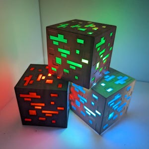 Puede incluir: Tres lámparas cúbicas estilo Minecraft apiladas. Las lámparas son de color gris oscuro con formas recortadas que brillan en rojo, verde y azul. Las lámparas están sobre una superficie blanca con un fondo degradado.