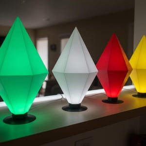 Puede incluir: Cuatro lámparas en forma de diamante en verde, blanco, rojo y amarillo. Cada lámpara tiene una base negra y un diseño geométrico. Las lámparas están dispuestas sobre una superficie clara.