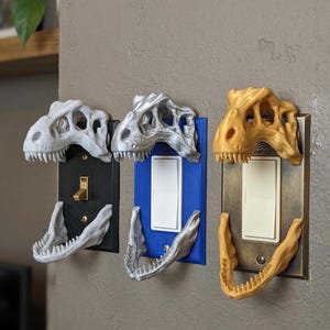May include: Three dinosaur skull light switch covers in white, silver, and gold. The covers are mounted on black, blue, and bronze switch plates. The jaws of the dinosaurs are open, as if they are biting the light switches.