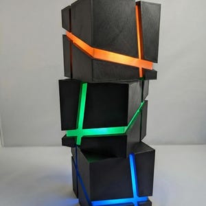 May include: A modern, geometric lamp composed of stacked black cubes. The lamp features illuminated cross-shaped accents in orange, green, and blue, creating a striking visual effect. The overall design is contemporary and artistic.