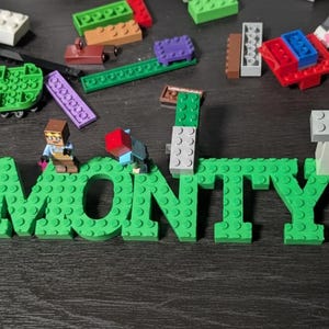 May include: Green LEGO blocks spelling out the name "MONTY". The letters are constructed with various LEGO bricks, and small LEGO figures are placed on top. Other LEGO pieces are scattered around.