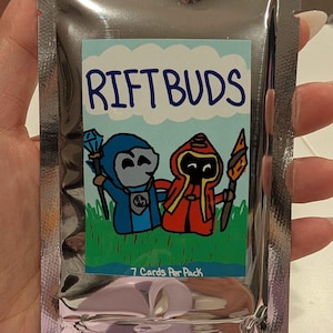 Puede incluir: Un paquete de papel de aluminio plateado que contiene un paquete de cartas con la etiqueta "RIFTBUDS". La etiqueta presenta personajes de dibujos animados con túnicas azules y rojas, sosteniendo bastones, sobre un fondo de cielo azul y hierba verde. También se ve el texto "7 Cards Per Pack".