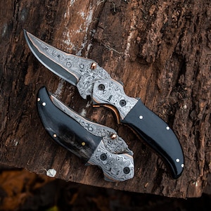 May include: A folding pocket knife with a Damascus steel blade featuring a swirling gray and silver pattern, paired with a smooth black handle accented by small brass pins. The knife is shown in both an open and a closed position for display.