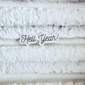 May include: A white sticker with the phrase "Hell Yeah!" in a cursive font. The sticker is placed on a textured, white surface with horizontal lines.