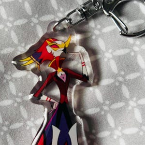 May include: A clear acrylic keychain featuring a cartoon character with red, yellow and white flames, a black and purple outfit, and a white star on their chest. The character is standing with one hand on their hip and the other raised to their chin.