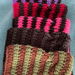 May include: A stack of five crocheted striped scarves in various colors, including purple, red, pink, black, and green.
