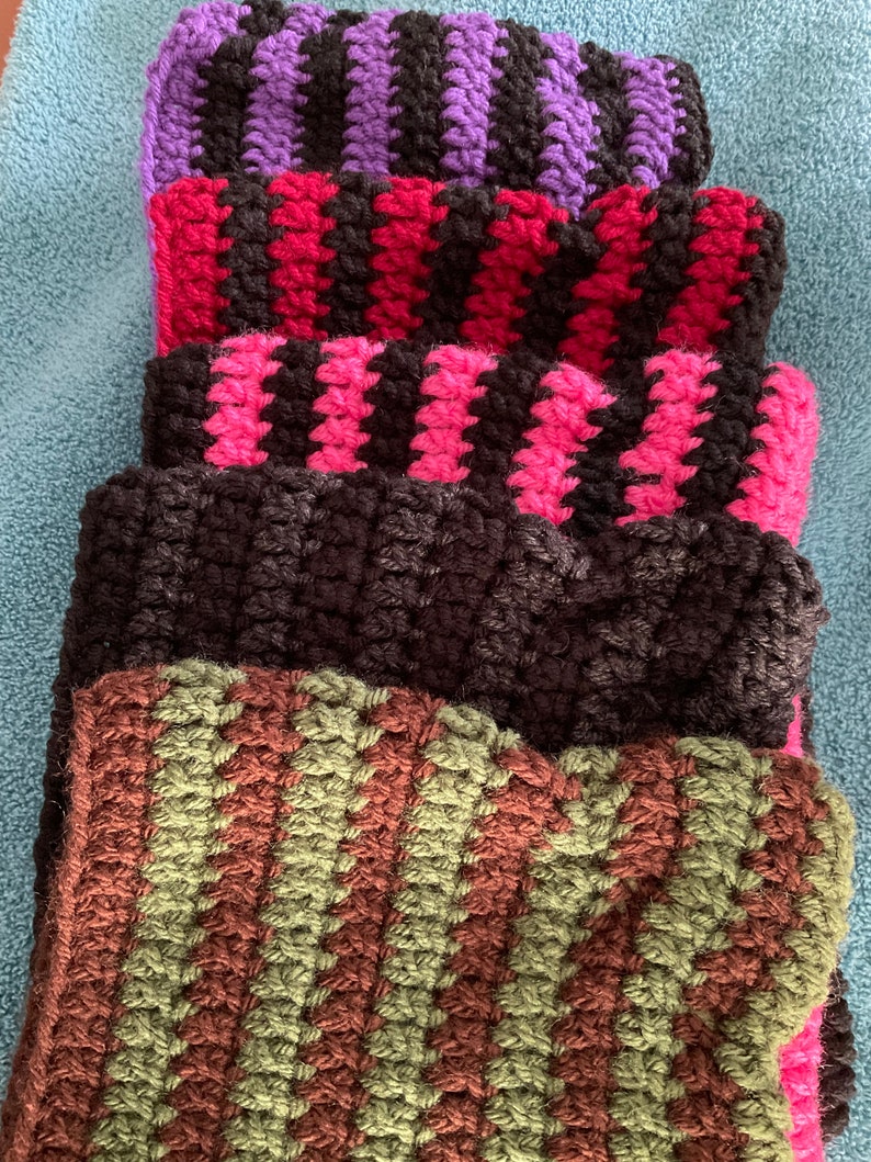 May include: A stack of five crocheted striped scarves in various colors, including purple, red, pink, black, and green.