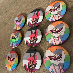 Pode incluir: Nove botões redondos com diferentes designs de bandeiras do orgulho. Cada botão apresenta um personagem de desenho animado com um fundo arco-íris, preto e branco ou rosa e branco.