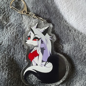 Puede incluir: Un llavero acrílico transparente con un personaje de lobo de dibujos animados. El lobo tiene pelaje blanco, una cola negra y detalles en rojo. El personaje lleva un top rojo y un collar negro. El llavero tiene un cierre metálico plateado.