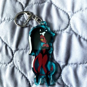 May include: A clear acrylic keychain featuring a cartoon character with red skin, black hair, and a devil tail. The character is wearing a black outfit with red fishnet stockings. The keychain has a silver metal clasp.
