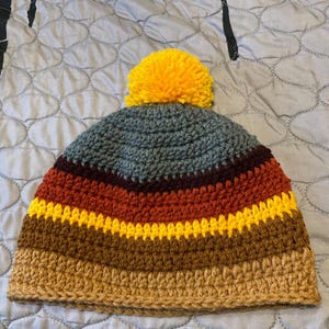 Può includere: Un cappello all'uncinetto fatto a mano con un pompon giallo. Il cappello presenta strisce orizzontali nei toni del marrone, arancione, giallo e grigio. Un accessorio caldo e accogliente per il clima freddo.