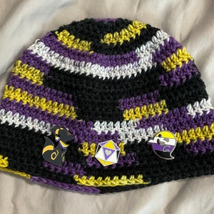 Könnte beinhalten: Eine schwarze, lila, weiße und gelbe Häkelmütze mit drei daran befestigten Anstecknadeln. Die Anstecknadeln sind ein schwarzer und gelber Comic-Charakter, ein lila und gelber Würfel und ein lila und gelber Geist.