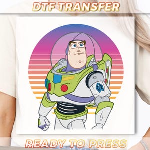 May include: A white DTF transfer featuring a cartoon illustration of Buzz Lightyear in his space suit. The design includes a retro sunset background with the text "READY TO PRESS". The words "DTF TRANSFER" are at the top.