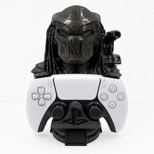 May include: A black 3D-printed bust of the Predator character featuring dreadlocks and a shoulder-mounted cannon, serving as a stand for a white and black wireless game controller with a PlayStation logo on the base support structure.