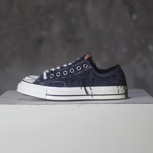 May include: A pair of dark blue denim sneakers with white soles and laces. The shoes have a distressed look with white paint-like accents on the sides. The sneakers are displayed on a white cube against a gray background.