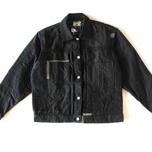 May include: A black long-sleeve jacket featuring a collared neckline, a front button closure with silver-toned buttons, and a textured patchwork design. The garment includes a chest pocket and visible white stitching details on the fabric panels.