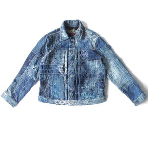 Pode incluir: Casaco de ganga azul com design de patchwork. O casaco tem uma frente com botões, mangas compridas e gola. O patchwork inclui vários tons de azul e costuras brancas.