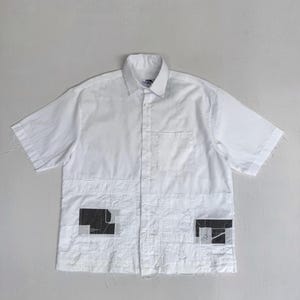 Pode incluir: Camisa branca de manga curta com botões e gola. A camisa apresenta patches com desenhos geométricos em preto e branco na parte inferior da frente. As bordas da camisa têm um aspecto desgastado e inacabado.