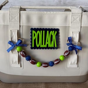 May include: A beige tote bag featuring a rectangular black plaque with the name POLLACK in bright green letters. A decorative beaded garland hangs from the bag's side rings, consisting of brown football-shaped beads, lime green beads, and blue glitter beads, accented with blue and white polka-dot ribbon bows.