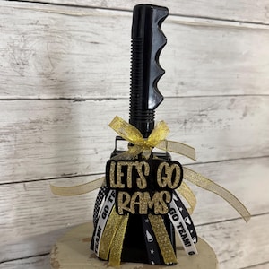 May include: A black plastic noisemaker handle with a vertical ribbed grip. A gold glitter cutout reads "LET'S GO RAMS" in a bold font. The base is decorated with multiple ribbons in gold, black, and white, featuring the text "GO TEAM!" and star icons.
