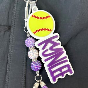 May include: A softball-themed keychain with a yellow softball design, red stitching, and the name "Kayne" in purple. The keychain includes purple and silver beaded accents and a purple tassel.