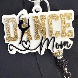May include: A white and gold glitter acrylic Dance Mom badge reel. The badge reel features the words "DANCE Mom" with a dancer silhouette. The badge reel has a black and silver beaded accent.