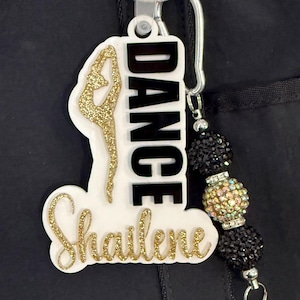 May include: A white and gold glitter acrylic keychain with the word "DANCE" in black block letters. The keychain features a dancer silhouette and the name "Shailene" in gold glitter script. A beaded charm with black and iridescent beads is attached.