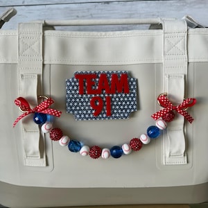May include: A beige cooler bag decorated with a rectangular patch featuring a blue and white star pattern and the red text TEAM 91. A beaded garland with red, white, and blue beads, including baseball-themed beads, hangs from two red polka-dot bows.